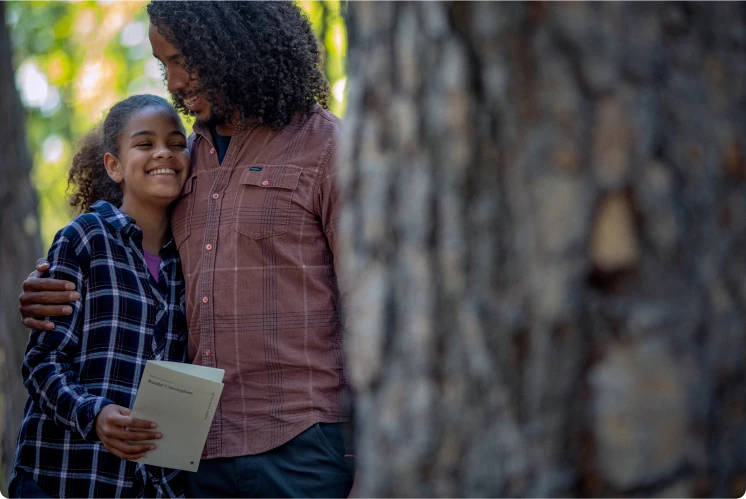a brother and sister in background hugging and smiling with a blurred memorial tree in the foreground