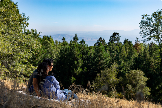 A couple sits quietly on a hillside overlooking a forested valley under a clear blue sky.